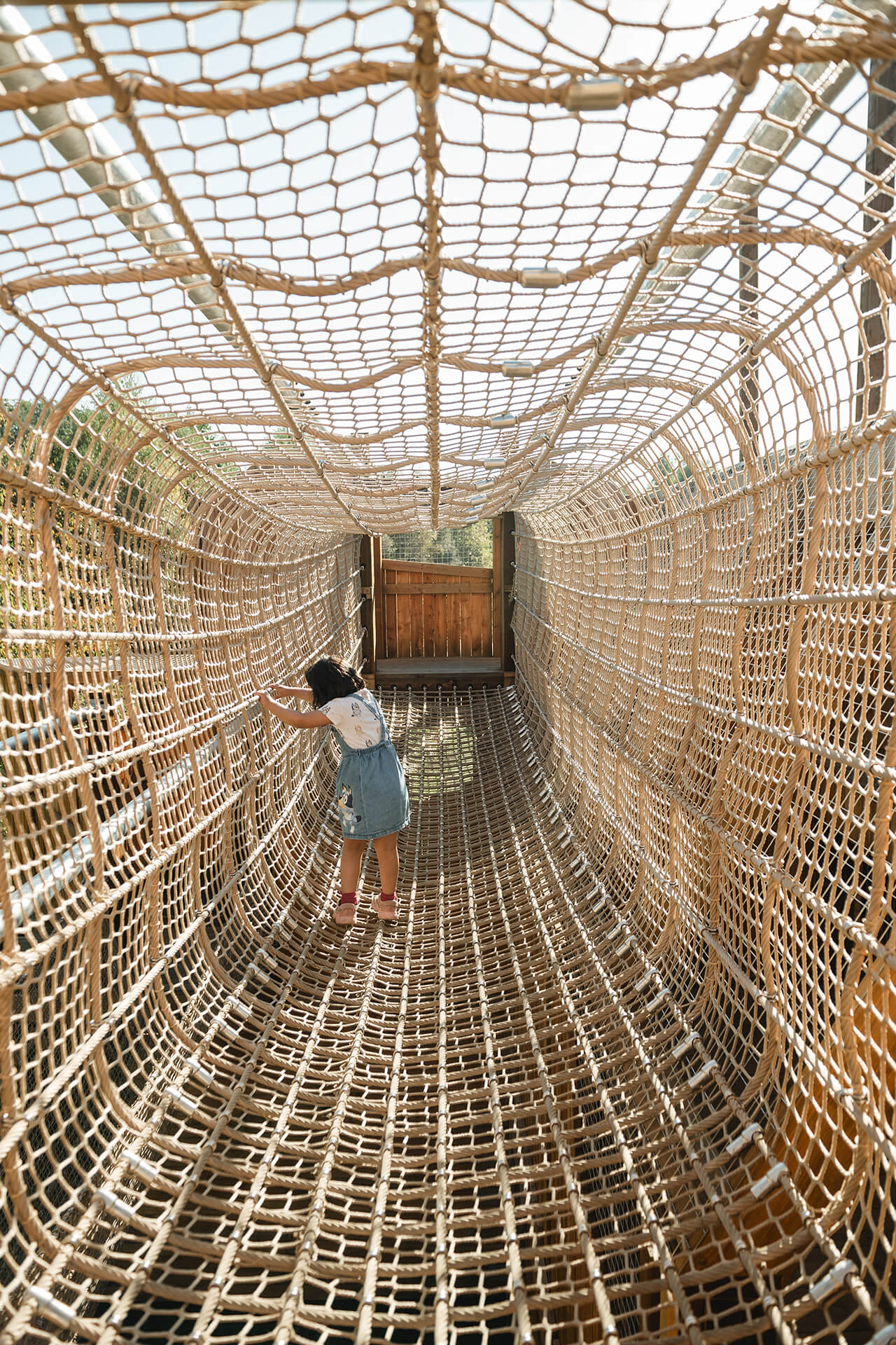 Image of child walking across a fully enclosed rope netting bridge built by CAP.CO adventure playground at Almond Valley photography by Lindsey Mack