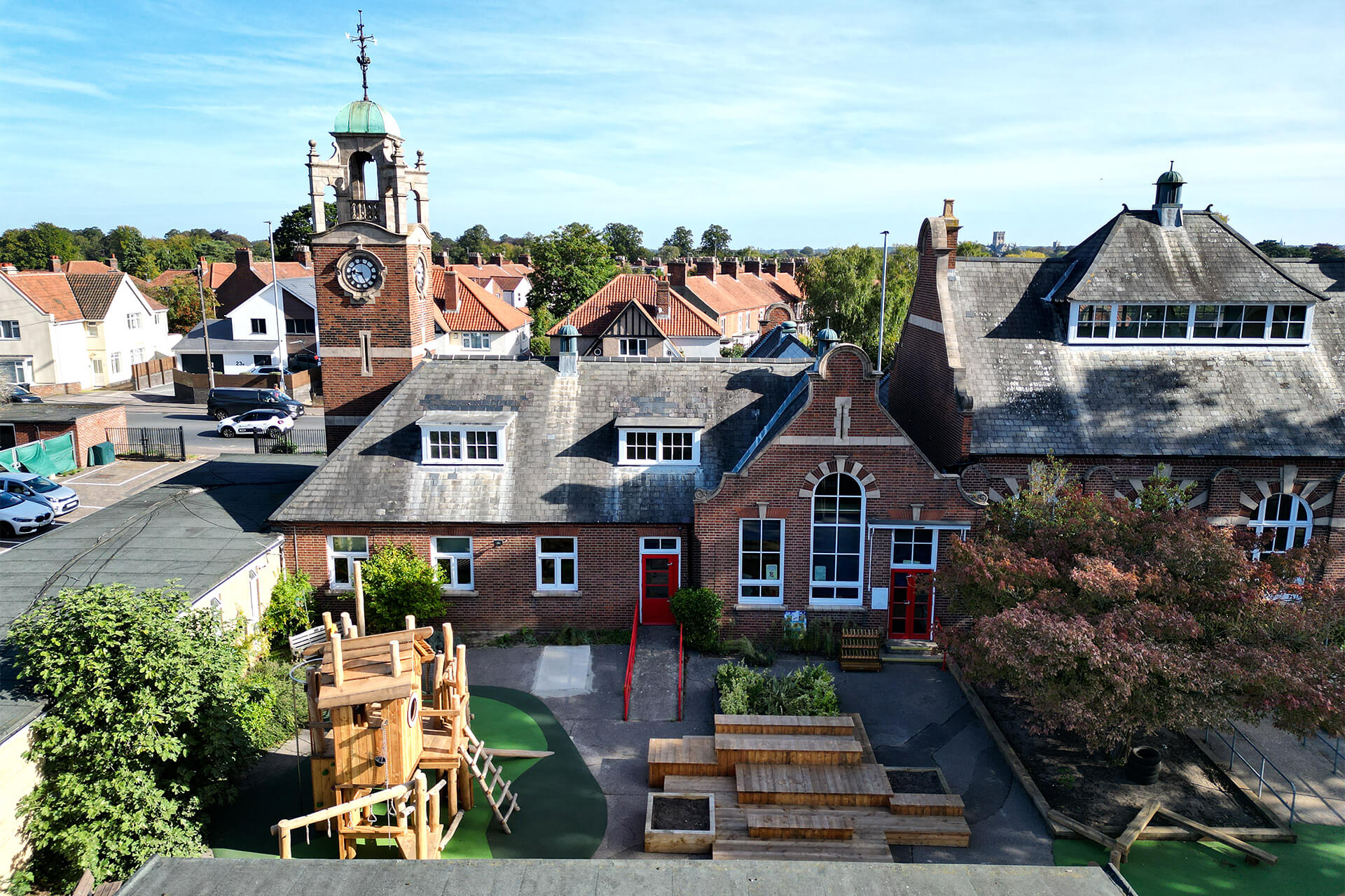 Aerial view of the play tower and stacked outdoor classroom decks in front of the Colman Infant School clock tower and red brick building, designed and built by CAP.CO. Photography by George Browne