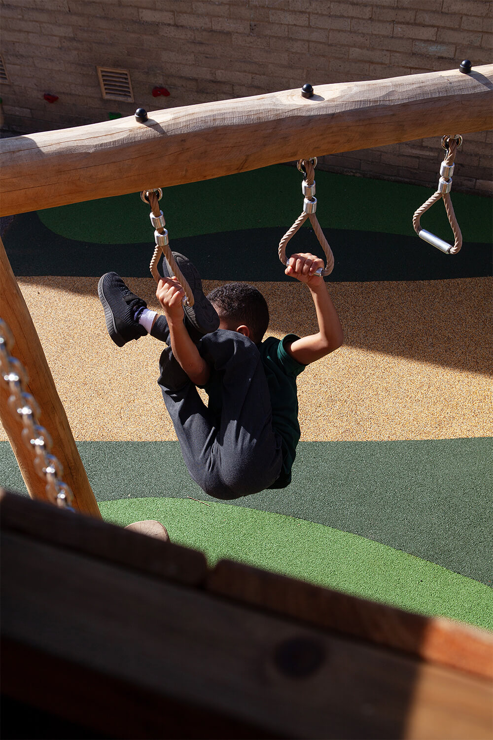 Child hanging from trapeze rings above colourful rubber crumb safety surfacing at Colman Infant School, built by CAP.CO. Photography by George Browne