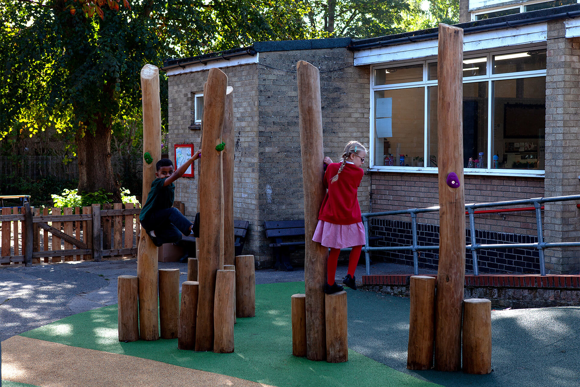 Children climbing tall robinia poles with climbing holds in the ground play area at Colman Infant School, designed by CAP.CO. Photography by George Browne