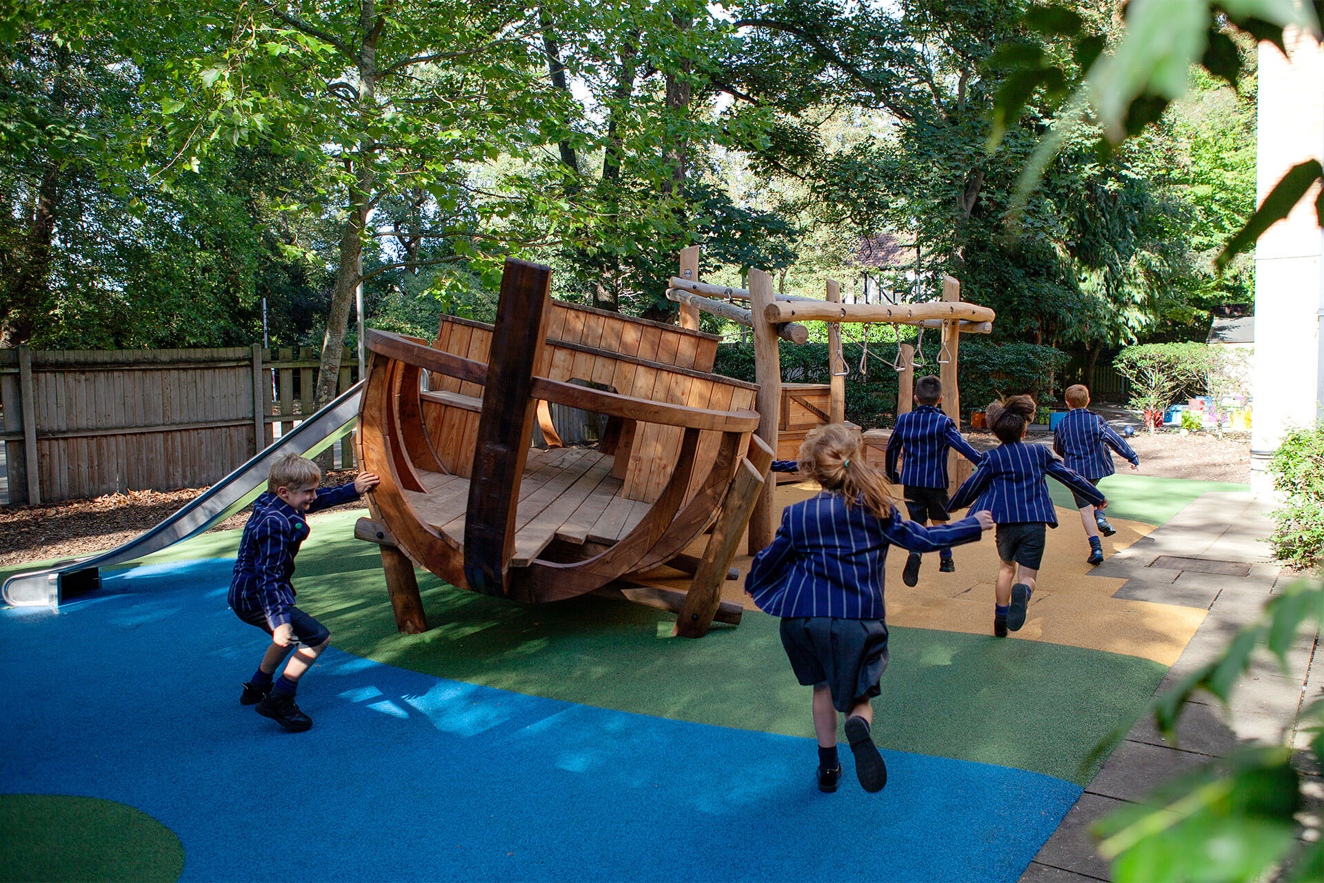 Children running towards the wherry boat and slide across blue rubber crumb surfacing at Town Close School, Norwich, a school playground by CAP.CO. Photography by George Browne