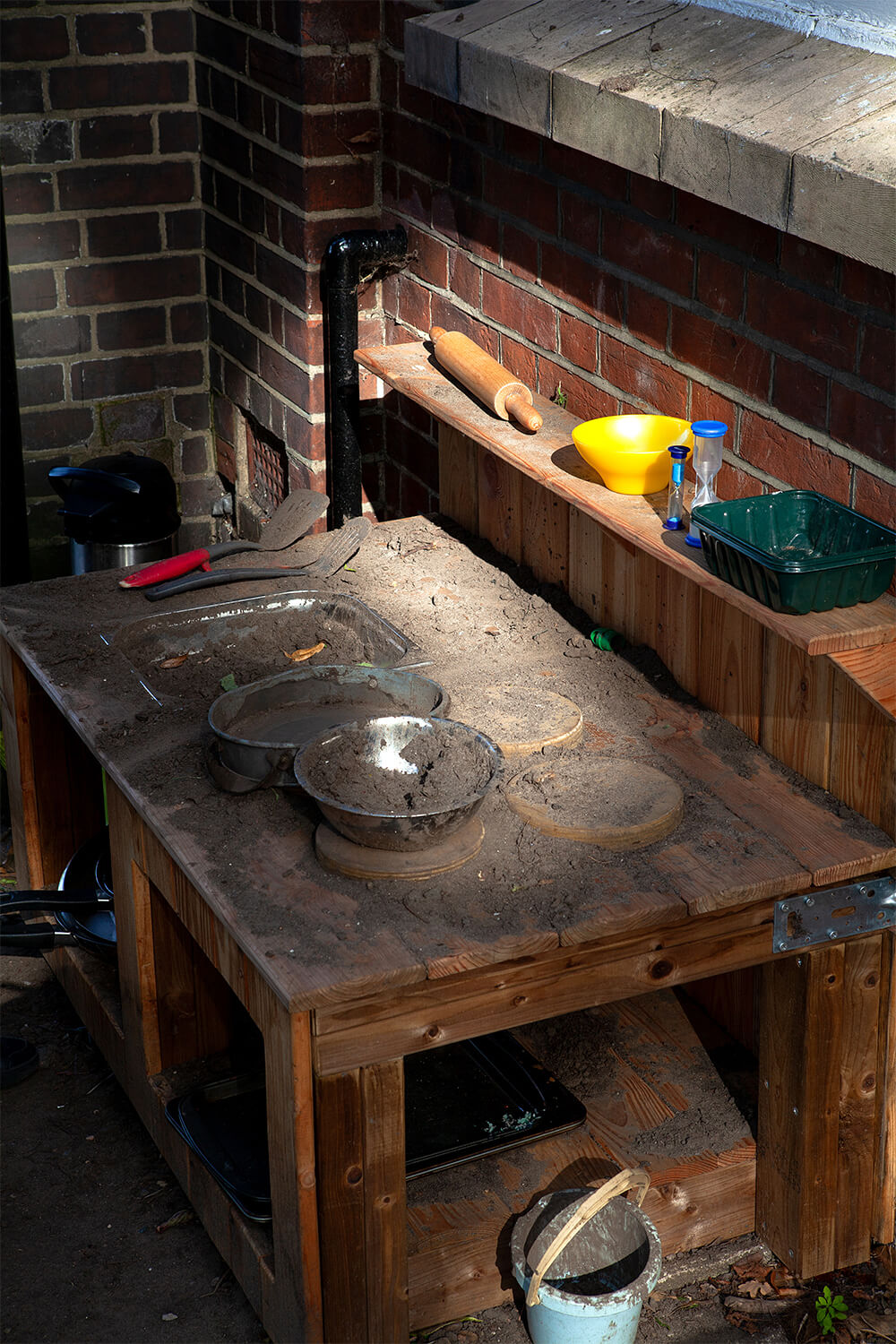 Wooden mud kitchen with pans, bowls and utensils against the school’s red brick wall at Colman Infant School, built by CAP.CO. Photography by George Browne
